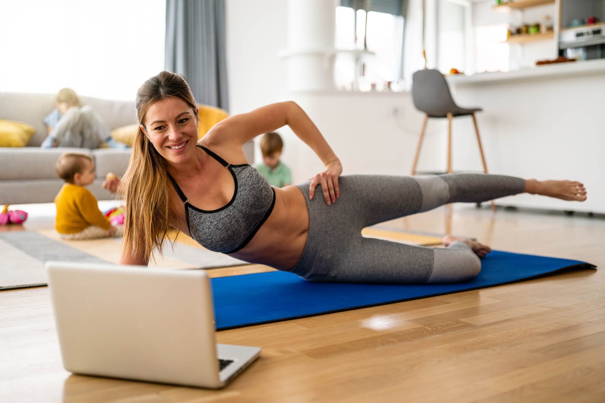 Young woman, mother exercising at home in living room, father playing with kids in background